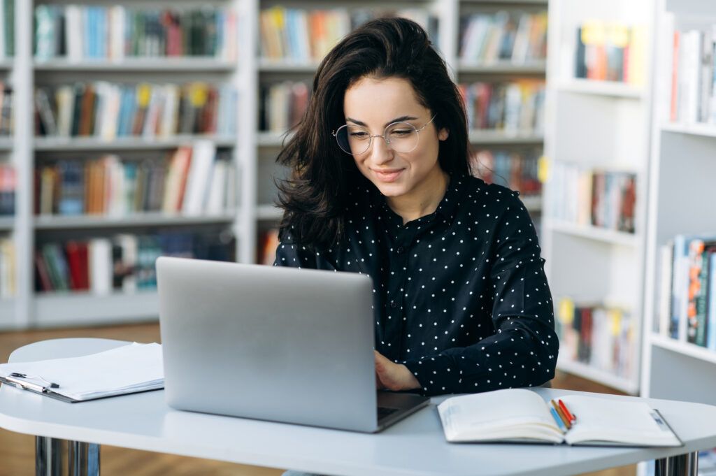 An educator in round glasses and a polka-dot blouse smiles at her laptop at a library table, an open notebook and colored pencils beside her