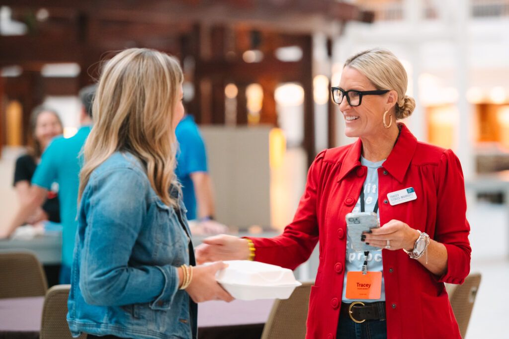 engage2learn consultant Tracey Stokes-Marriott, in a red jacket and glasses, smiles as she greets a partner in a denim jacket at an event
