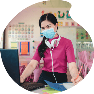 Teacher with mask on looking at laptop in colorful classroom
